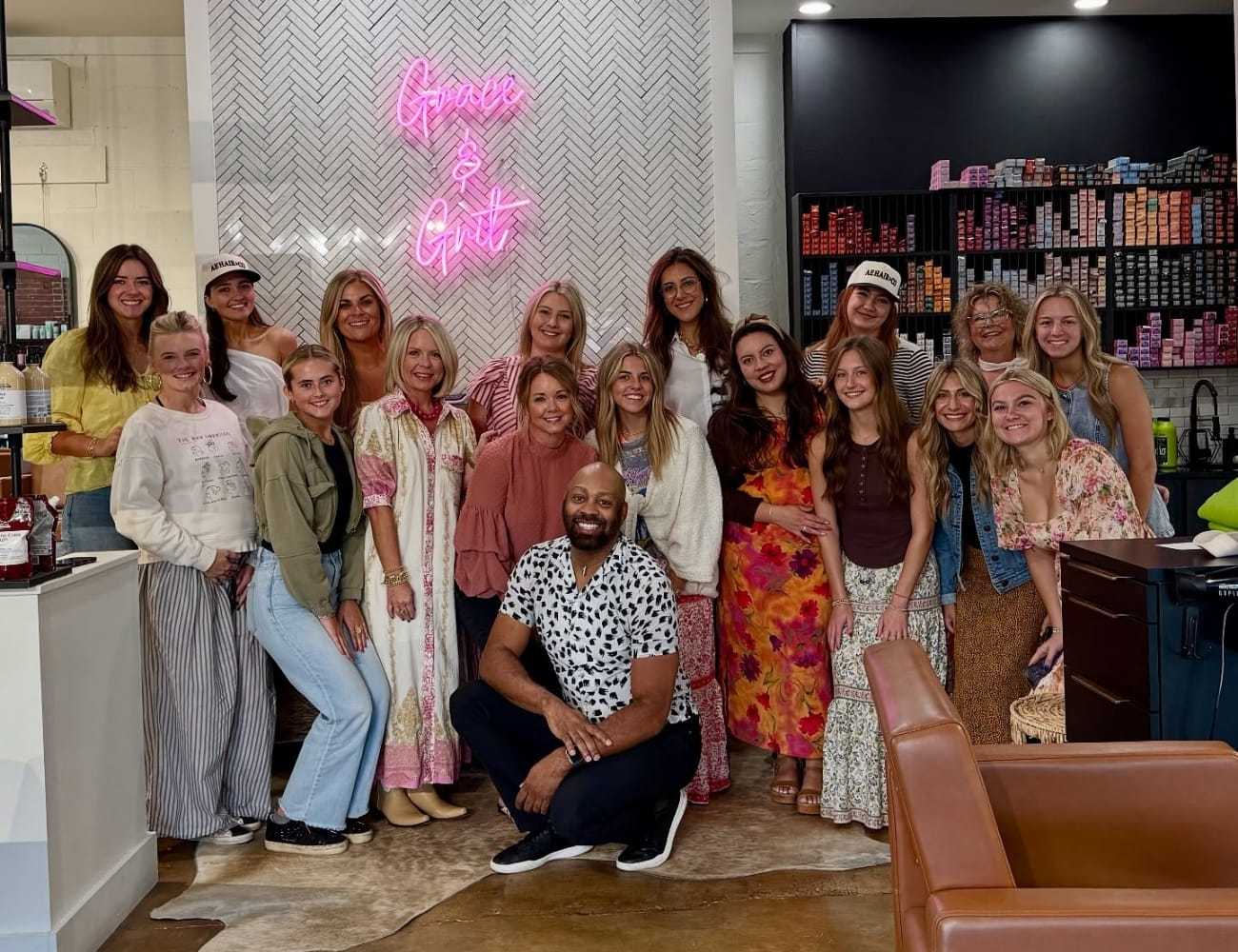 Group of people smiling in a hair salon with a neon "Grace & Grit" sign in the background.